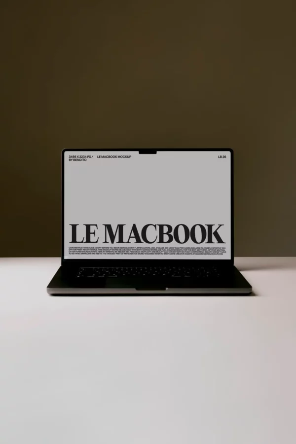 A black laptop is open on a white surface with a gray screen displaying large text that reads LE MACBOOK. The background is plain and dark brown.