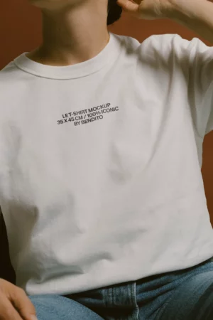 A person wearing a plain white t-shirt with black text that reads LET-SHIRT MOCKUP 35 X 45 CM / 100% ICONIC BY BENDITO. The person is seated and partially cropped, with one hand near their neck.