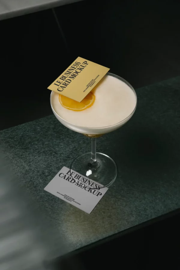 A cocktail in a coupe glass garnished with an orange slice and a yellow business card; another business card lies on the green countertop nearby.
