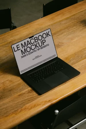 A black MacBook laptop displaying a screen with the text LE MACBOOK MOCKUP 3456 X 2234 PX / BY BENDITO sits open on a wooden table surrounded by black chairs.