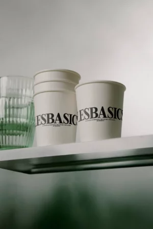 Two white paper cups with bold black text reading LES BASICS are stacked on a glass shelf beside clear, ribbed glass cups against a blurred, neutral background.