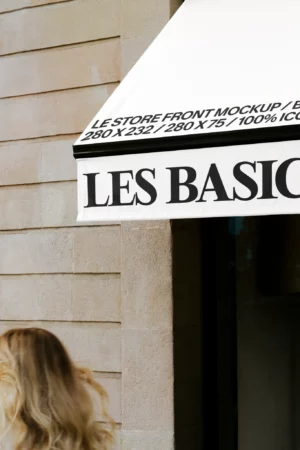 A white store awning with black text reads LES BASICS, with mockup dimensions and details printed above. Part of a stone wall and a person with long blonde hair are visible in the foreground.
