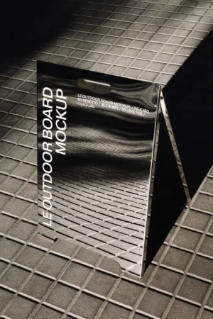 A reflective A-frame outdoor board stands on a tiled surface, displaying the text LE OUTDOOR BOARD MOCKUP in bold white letters. The board captures the shadows and grid pattern of the pavement around it.