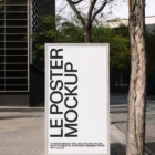 A large vertical poster mockup stands on a city sidewalk near a tree, displaying bold black text reading “LE POSTER MOCKUP” on a white background, with smaller text at the bottom. Buildings and trees are in the background.