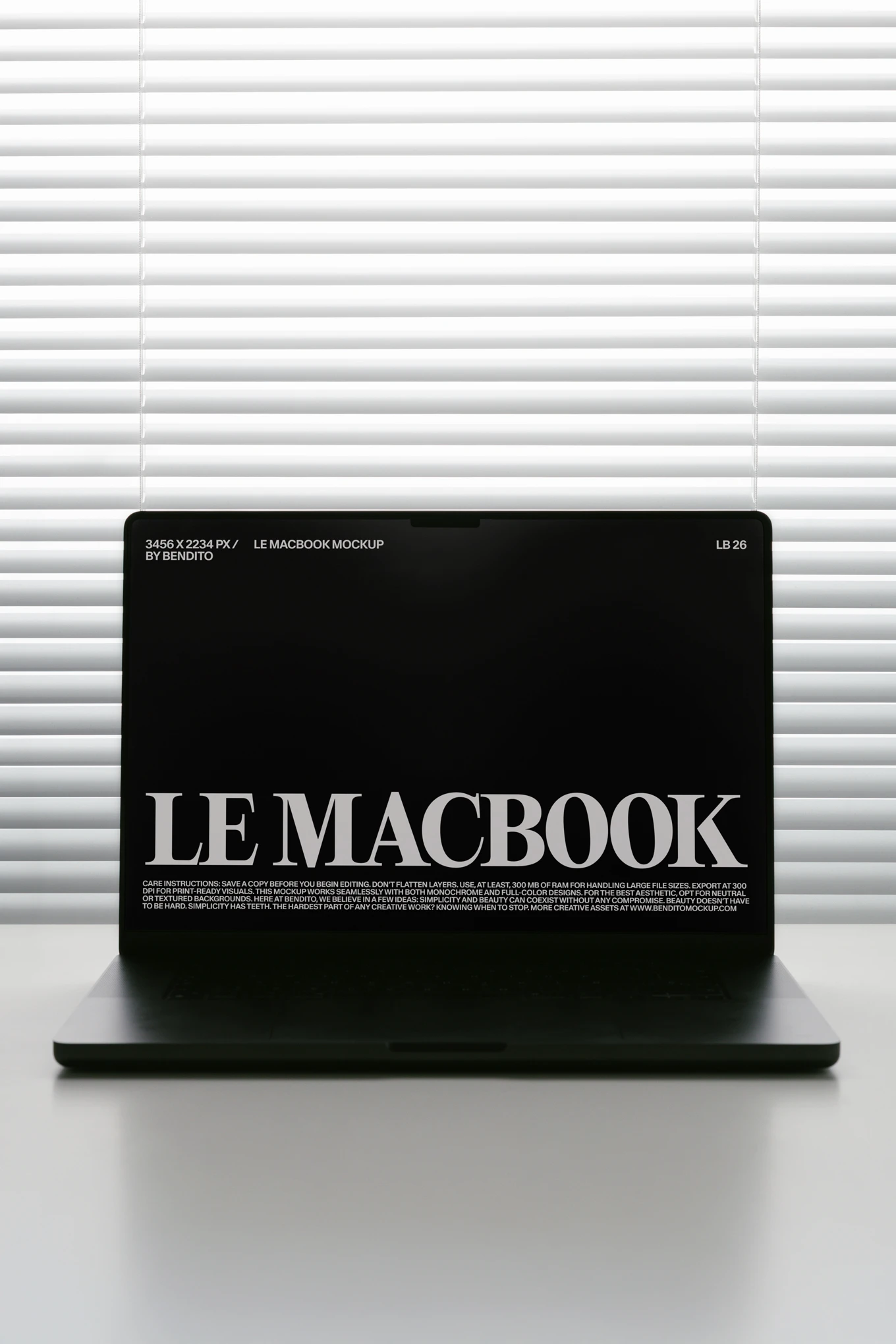 A dark laptop sits on a white desk in front of closed window blinds, displaying a black screen with bold white text reading LE MACBOOK.