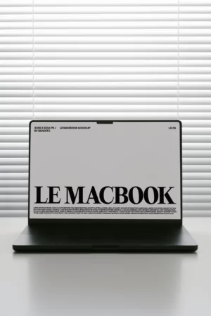 A black laptop displaying a screen with the text LE MACBOOK in bold, sits on a white surface in front of closed white blinds.