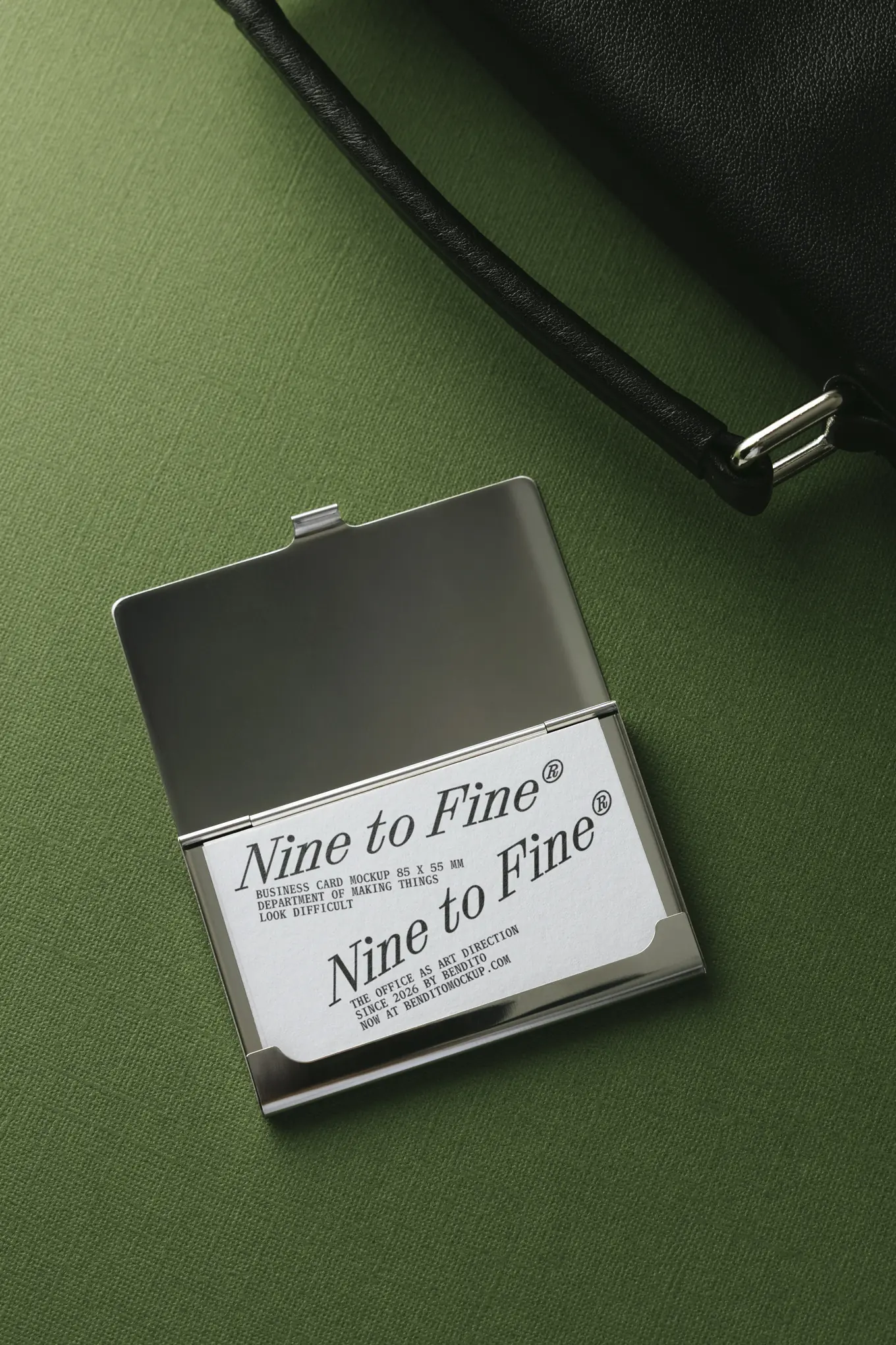 A silver business card holder sits open on a green textured surface, displaying a card that reads Nine to Fine. Part of a black handbag is visible in the upper right corner.