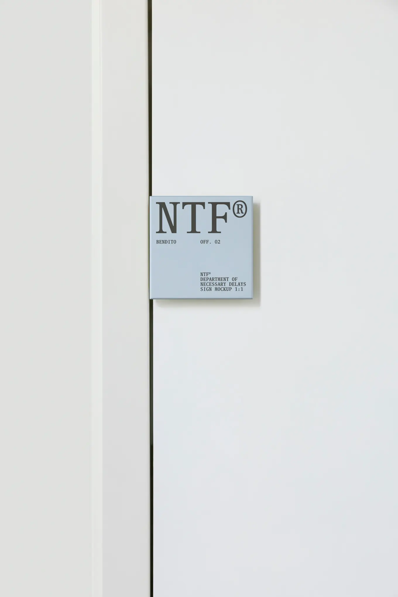 A small gray sign with the large letters NTF® and smaller text below is mounted on a white wall next to a white door or panel. The design is minimal and modern.