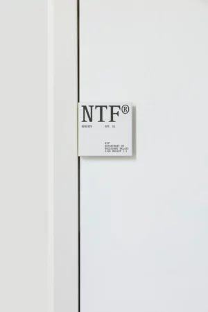 A white sign with large gray letters NTF® and smaller text below is mounted on a plain white wall next to a thin vertical gap or door frame.