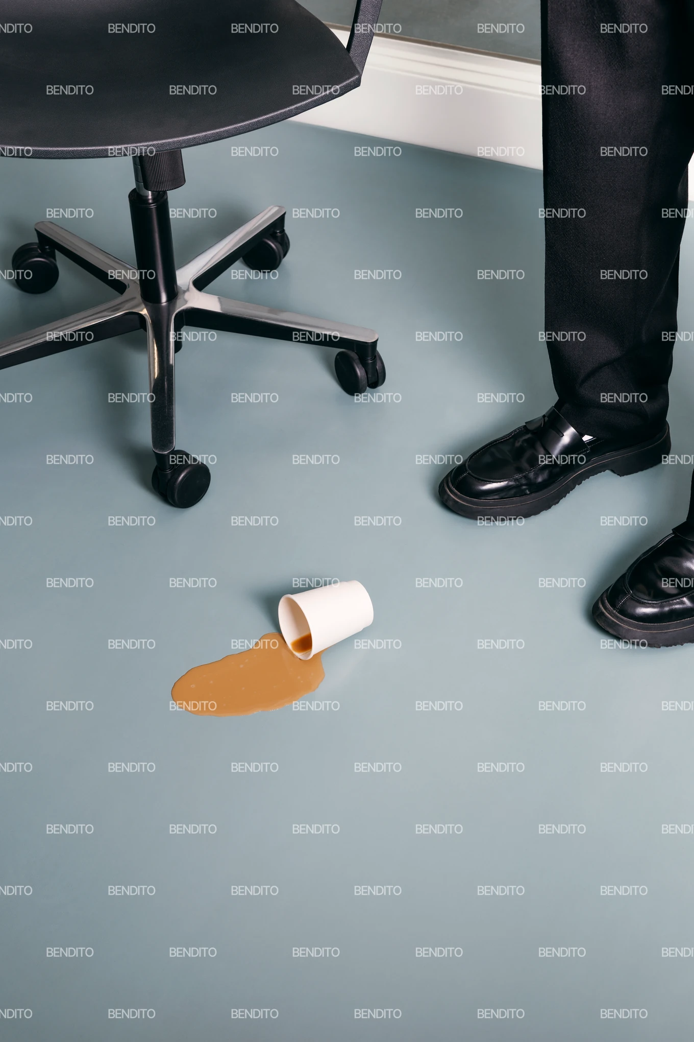 A white paper cup with spilled coffee lies on the floor near a rolling office chair and a persons polished black shoes.