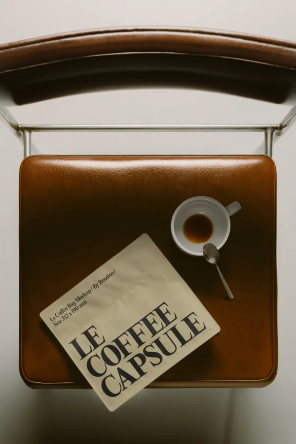 A top-down view of a brown leather chair with a white cup holding a small amount of coffee, a spoon, and a beige packet labeled LE COFFEE CAPSULE on the seat.
