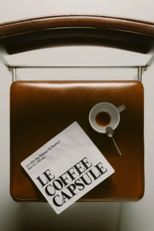 A top-down view of a brown leather chair with a white cup of espresso, a spoon, and a white packet labeled LE COFFEE CAPSULE resting on the seat.