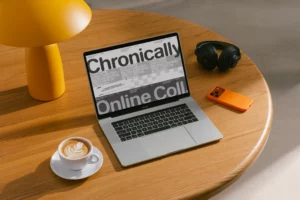 A laptop displaying bold text, “Chronically Online Coll,” sits on a wooden table next to a yellow lamp, black headphones, an orange smartphone, and a cup of coffee with latte art.