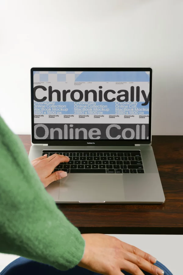 A person in a green sweater uses a laptop on a wooden table. The laptop screen displays large, bold text that reads Chronically Online Coll. with other smaller overlapping text in blue and white.