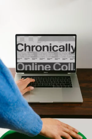 A person uses a laptop on a wooden desk. The laptop screen displays large text that reads Chronically Online Coll. The persons hand rests on the keyboard.