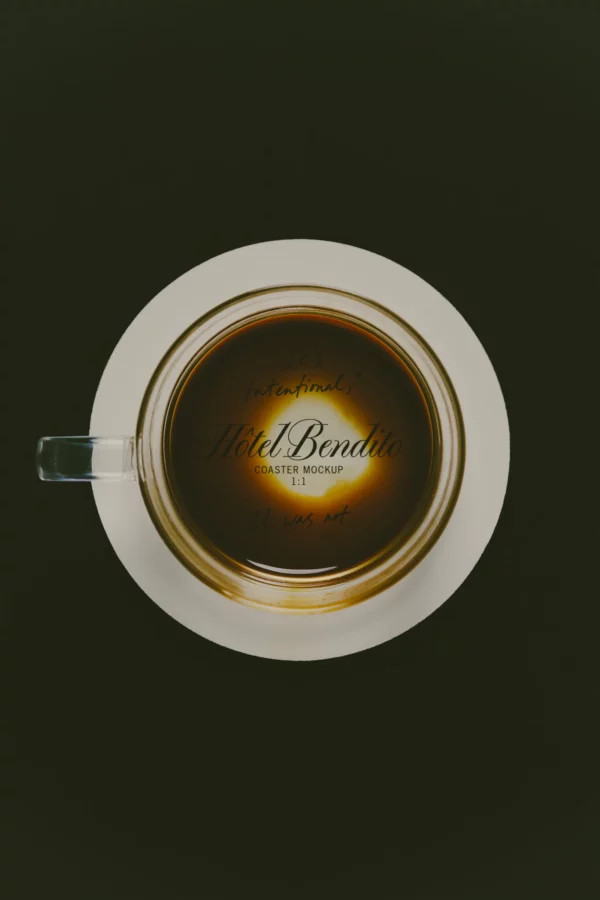 Round coaster mockup printed Hôtel Bendito, shot from directly above and seen through a glass cup of dark coffee resting on it, its pale rim showing beyond the base of the glass, on a deep olive green background.