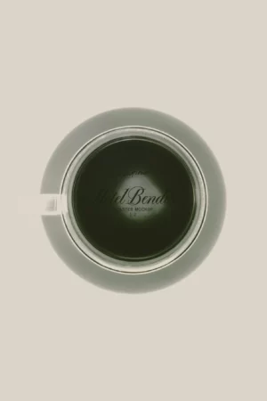 Round coaster mockup printed Hôtel Bendito, shot from directly above and seen through a glass cup of dark coffee resting on it, its pale rim showing beyond the base of the glass.